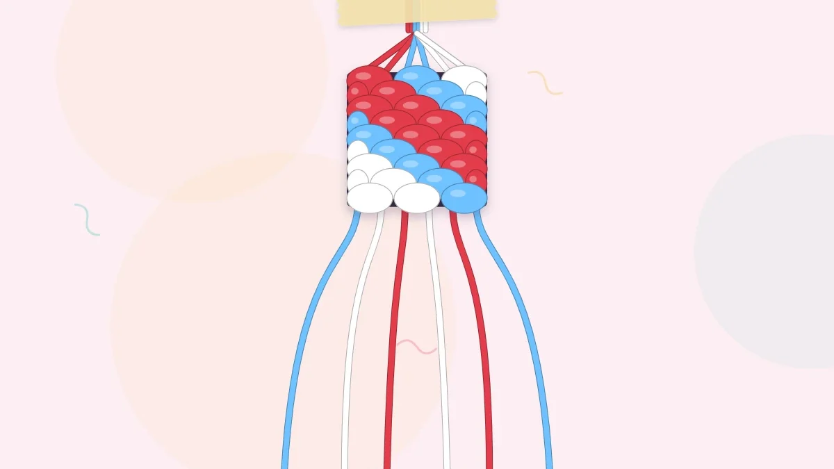 A candy stripe bracelet in red, sky blue and white being knotted, taped at the top with loose strings fanned out below