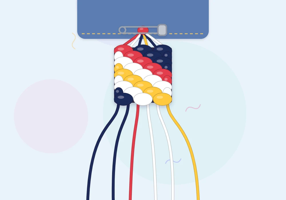 A candy stripe bracelet in progress held by a safety pin, with red, white, navy and yellow diagonal stripes